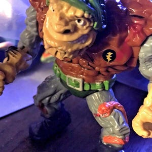 May include: A vintage action figure of a mutant character with a tan face, green head covering, and brown and orange body armor. The figure has gray arms and legs, a green belt, and a red snake wrapped around its leg. A black emblem with a lightning bolt is on the chest.