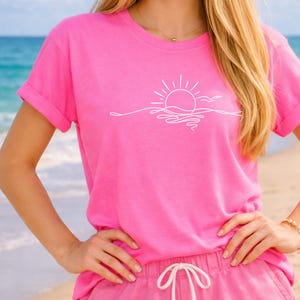 Puede incluir: Una camiseta rosa brillante con un dibujo lineal blanco de una puesta de sol sobre el océano. La camiseta de manga corta tiene las mangas enrolladas y se combina con pantalones cortos rosas. El diseño presenta un sol, olas y un pájaro.