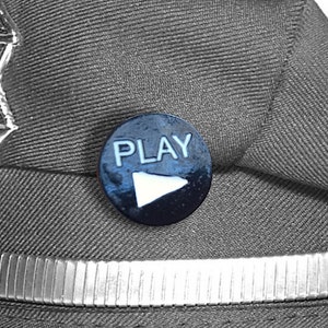Könnte beinhalten: Ein schwarzer und silberner Polizeihut mit einem silbernen Abzeichen, auf dem "Patrolman" steht, und einem blauen Knopf mit dem Wort "PLAY" und einem weißen Play-Symbol.