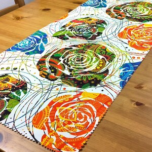 以下が含まれることがあります： 青、緑、オレンジ、黄色の色合いで、繰り返しの花柄が特徴的なカラフルなテーブルランナー。 白い背景に、複雑なディテールを持つ様式化されたバラのデザイン。 木製のテーブルの上に置かれています。