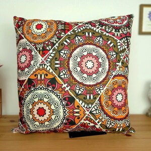 Puede incluir: Una almohada decorativa con un patrón geométrico que presenta colores rojo, naranja, marrón y blanco. El patrón está formado por círculos y espirales repetidos.
