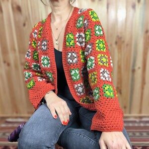 May include: A red crocheted cardigan with a granny square pattern in green, brown, and white. The cardigan has a V-neck and long sleeves.