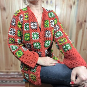 May include: A red, green, and brown crocheted cardigan with a granny square pattern. The cardigan is open in the front and has long sleeves.