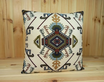 Puede incluir: Una almohada blanca con un patrón geométrico en tonos de azul, verde, marrón y rojo. El patrón presenta una forma de diamante central con detalles intrincados.