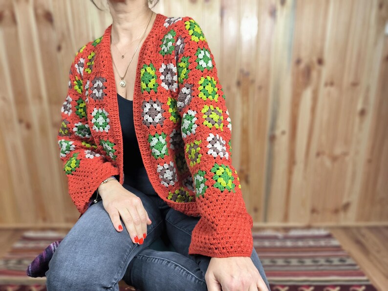 May include: A red crocheted cardigan with a granny square pattern in green, brown, and white. The cardigan has a V-neck and long sleeves.