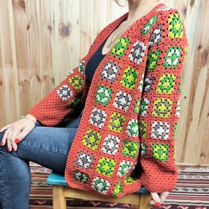 May include: A red, green, and brown crochet cardigan with a granny square pattern. The cardigan is open in the front and has long sleeves.
