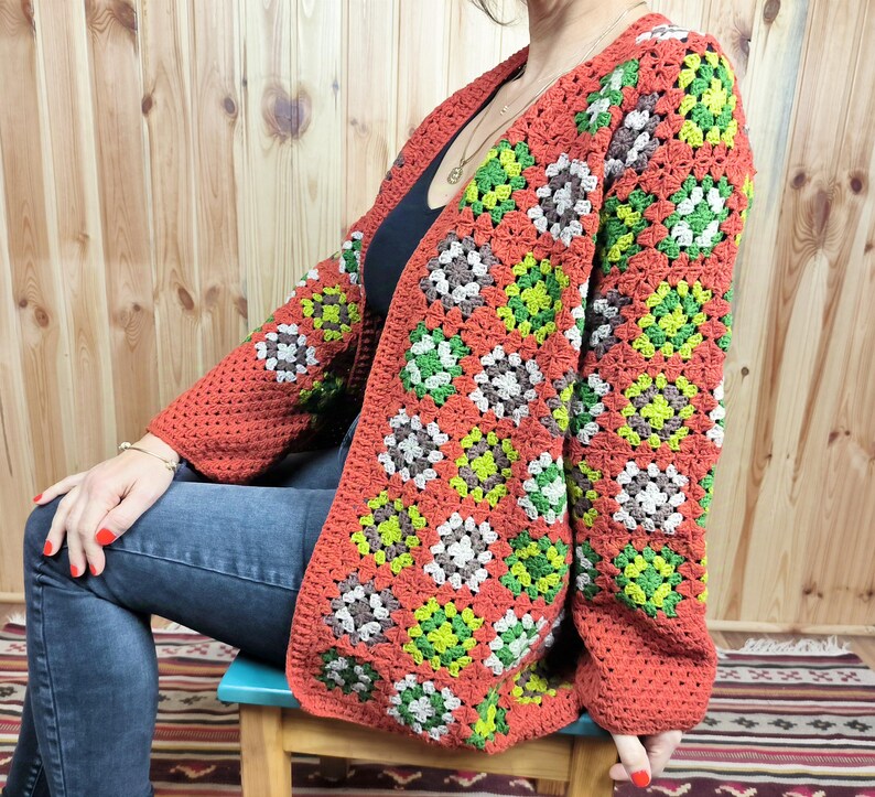 May include: A red, green, and brown crochet cardigan with a granny square pattern. The cardigan is open in the front and has long sleeves.