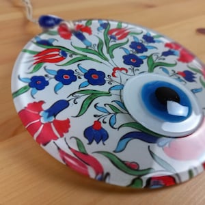 Könnte beinhalten: Runder Glasanhänger mit weißem Hintergrund und einem blauen bösen Auge. Der Anhänger ist mit roten, blauen und grünen Blumenmustern verziert.