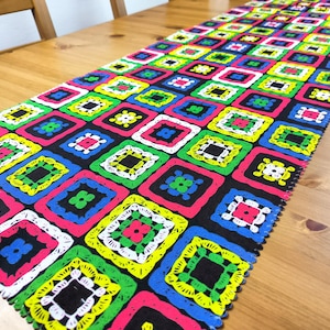 Può includere: Un runner da tavolo colorato con un motivo geometrico di quadrati in tonalità di verde, giallo, rosa, blu e bianco su uno sfondo nero.