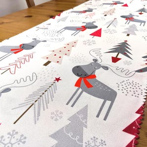 May include: White table runner with a red and gray Christmas design. The design features moose, Christmas trees, snowflakes, and stars.
