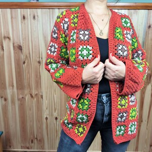 May include: A red, green, and brown crochet cardigan with a square pattern. The cardigan is open in the front and has long sleeves.
