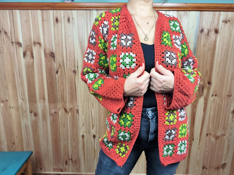 May include: A red, green, and brown crochet cardigan with a square pattern. The cardigan is open in the front and has long sleeves.
