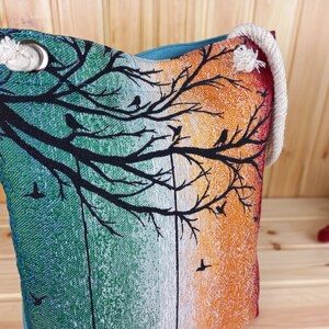 May include: A teal, white, and orange tote bag with a black silhouette of a tree with birds on the branches. The bag has a rope handle.