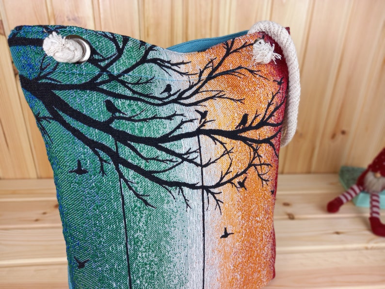 May include: A teal, white, and orange tote bag with a black silhouette of a tree with birds on the branches. The bag has a rope handle.