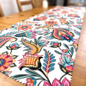 Peut inclure: Un chemin de table blanc avec un motif paisley coloré composé de motifs floraux roses, bleus, jaunes et verts.