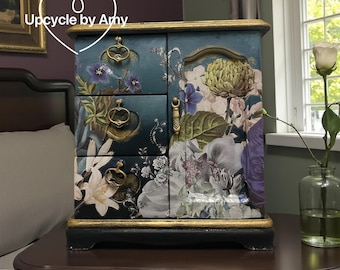 Op de afbeelding: Een tealkleurige sieradendoos met bloemmotieven en gouden accenten. De doos heeft drie lades en een kastdeur, allemaal versierd met botanische illustraties in paars, wit en groen. De tekst "Upcycle by Amy" staat in een hartvorm.