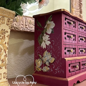 May include: A small, wooden jewelry box with multiple drawers. The box is painted a deep burgundy color and features a floral design with gold accents. The text "Upcycle by Amy" is visible on the bottom of the image.