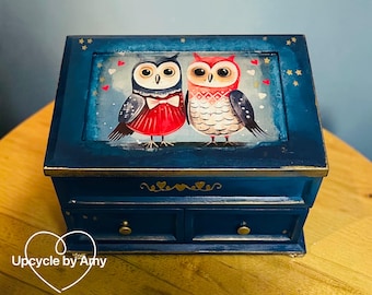 Op de afbeelding: Een donkerblauw houten sieradendoosje met een geschilderde illustratie van twee uilen op het deksel, één rood en één wit, omringd door kleine hartjes en sterren. De doos heeft twee kleine lades met gouden knoppen en hartjes.
