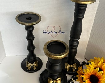 Op de afbeelding: Drie zwarte kaarsenstandaards met gouden accenten en gouden strikken. De kaarsenstandaards staan op een wit oppervlak met een zwarte en gouden kralenketting en een gele zonnebloem.