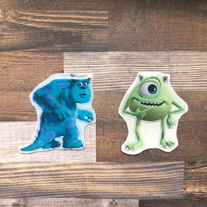 May include: Two cartoon characters from the movie Monsters Inc.  The blue character, Sully, is standing on the left. The green character, Mike, is standing on the right. Both characters are smiling.