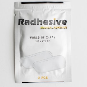 May include: A silver pouch with a black border containing two white medical adhesive patches. The pouch has the text "Radhesive Medical Adhesive" and "World of X-Ray Signature" printed on it. The pouch also has the text "2 PCS" printed on it.