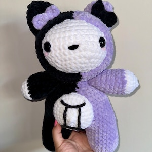 May include: A crocheted plush toy bear with a black and purple color scheme. The bear has a white face with black eyes and a black nose. The bear's belly is white with a black Gemini zodiac symbol.