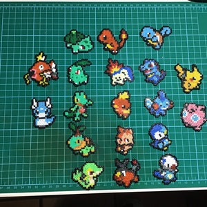 Könnte beinhalten: Eine grüne Schneidematte mit 20 bunten Pixel-Art-Pokémon-Charakteren aus Perlen. Die Charaktere umfassen Bisasam, Glumanda, Schiggy, Pikachu und mehr.