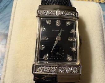 May include: A vintage Hamilton wristwatch with a rectangular black face and diamond accents. The watch features a black leather band and is set on a cream-colored surface. The watch face has the word "Hamilton" printed on it.