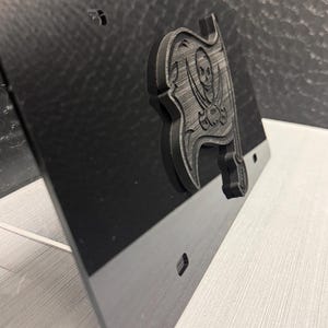 May include: Black license plate featuring a gray, 3D-printed pirate flag emblem with a skull and crossed swords. The plate has pre-drilled holes for mounting and a glossy finish. The design is a sports-themed accessory.
