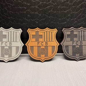 May include: Three wooden FC Barcelona crests in different colors: gray, brown, and black. Each crest features the team's logo, including the letters "FCB" and a soccer ball.