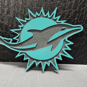 May include: A teal and gray emblem featuring a dolphin leaping through a sunburst design. The emblem has a textured surface and is set against a dark, textured background. The design is a sports team logo.