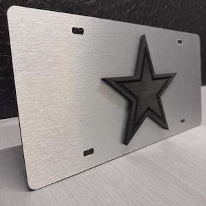 May include: Silver-colored license plate with a raised black star emblem. The plate has a brushed metal finish and four mounting holes. The star is a five-pointed design with a black outline.