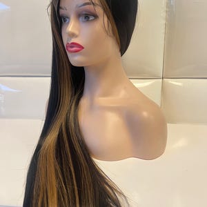May include: A long, black wig with blonde highlights is displayed on a mannequin head. The wig features a lace front and straight, sleek strands. The mannequin has a fair complexion, red lipstick, and is positioned against a white background.