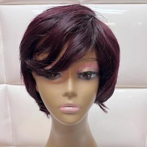 May include: A short, burgundy wig with layered bangs styled on a mannequin head. The wig has a natural-looking hairline and a slightly tousled texture. The mannequin has makeup applied, including pink eyeshadow and lipstick.