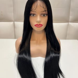 May include: A black wig with long, straight hair is displayed on a mannequin head. The wig features a natural-looking hairline and is styled with a center part. The mannequin has a fair complexion, red lipstick, and is set against a white background.