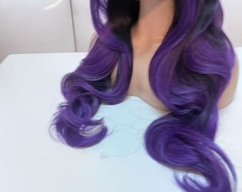 May include: A mannequin head displaying a long, wavy wig. The wig features a blend of black and vibrant purple, with the purple color concentrated towards the ends. The hair is styled with loose curls, creating a voluminous and eye-catching look.