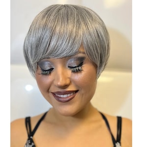 May include: A woman with short, silver-grey hair styled in a pixie cut. She is wearing makeup with silver eyeshadow and dark eyeliner.