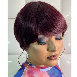 May include: A woman with short, burgundy-colored hair and gold hoop earrings. She is wearing colorful makeup and a blue and pink patterned shirt.