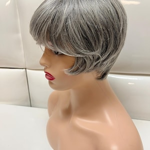 May include: A short, gray wig with a side part and bangs. The wig is styled in a layered bob with a slight wave.