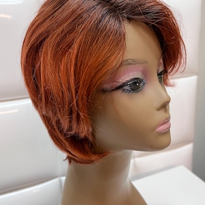 May include: A short, layered wig in shades of auburn and dark brown, styled on a mannequin head. The wig features a side part and a natural-looking hairline. The mannequin has makeup applied, including eyeshadow and lipstick.