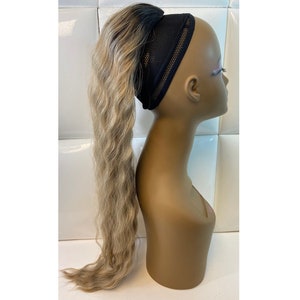 May include: A long, wavy blonde ponytail wig with a black headband is displayed on a mannequin head. The wig features dark roots that transition into a light blonde color. The mannequin is a neutral skin tone.