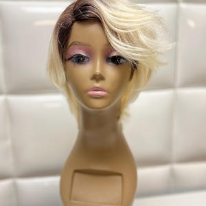 May include: A mannequin head with a short, layered wig. The wig features a dark brown root that fades into a light blonde. The mannequin has makeup applied, including pink eyeshadow and lipstick. The base of the mannequin is a light brown color.