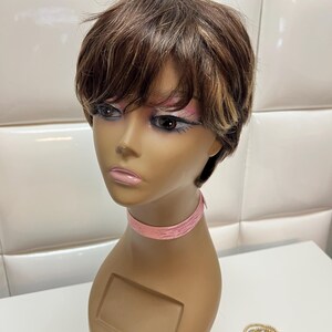 May include: A short, layered wig with brown and blonde highlights is displayed on a mannequin head. The mannequin has a pink ribbon around its neck and is set against a white background. A set of gold and silver rings are on the table.