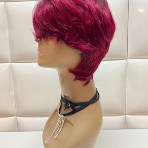 May include: A short, layered wig in a vibrant burgundy color, styled on a mannequin head. The wig features a dark root and a side-swept bang. A black choker with a rhinestone pendant is around the mannequin's neck.