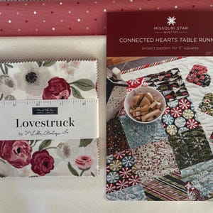 May include: A collection of quilting supplies, including fabric, a pattern, and a ruler. The fabric features floral patterns in shades of red, pink, and white. The pattern is for a "Connected Hearts Table Runner" and measures 20 x 38 inches.
