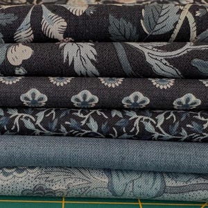 May include: A stack of fabric pieces in various shades of blue. The fabrics feature floral and botanical patterns in light blue and white, with a solid blue fabric at the bottom. The fabrics are stacked neatly, showcasing the different designs.