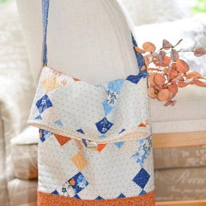 May include: A patchwork quilt bag with a blue and white floral pattern and a brown trim. The bag has a long blue strap and is hanging on a chair.