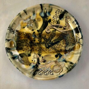May include: Round decorative plate with a unique design. The plate features a yellow, brown, and blue abstract pattern with gold-colored dragon and paisley accents. Black dot patterns and circular designs add detail.