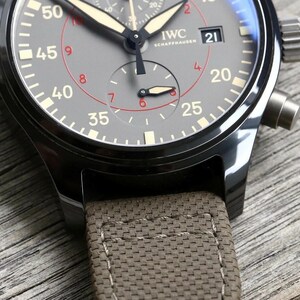 May include: An IWC Schaffhausen chronograph watch with a dark gray dial, featuring white and red numerals, luminous hands, and a date window at the three o'clock position. The watch has a black metal case and a textured, olive-colored fabric strap.