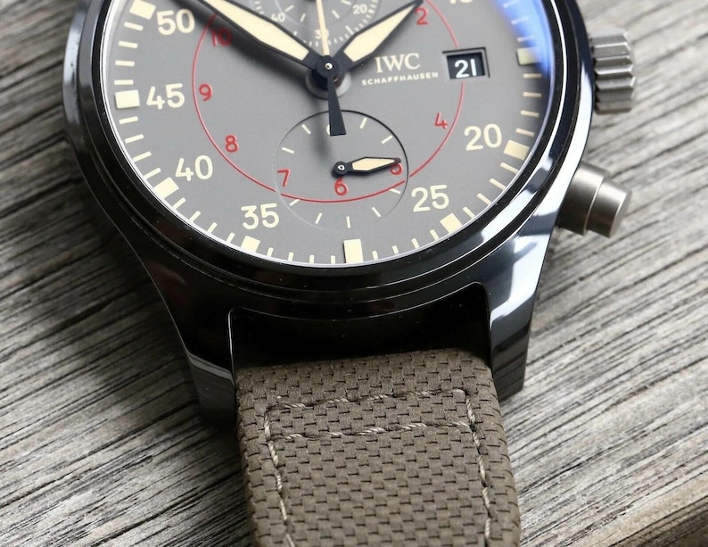 May include: An IWC Schaffhausen chronograph watch with a dark gray dial, featuring white and red numerals, luminous hands, and a date window at the three o'clock position. The watch has a black metal case and a textured, olive-colored fabric strap.